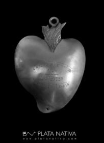 Exvoto en forma de corazón flamígero, símbolo del fervor y la fe, con orificio superior para colgar. Presenta una dedicatoria grabada "Los católicos meicanos a la milagrosa Virgen de Luján!, lo que indica su ofrenda colectiva como muestra de devoción y agradecimiento a la Virgen de Luján, patrona de la Argentina.. Alto 9 cm.